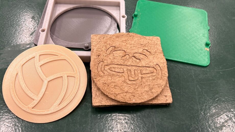 Several components of a 3D printed coaster mold laid out on a table, including a beige 3D printed pattern, a pressed cardboard coaster with an embossed face design and the mold parts.