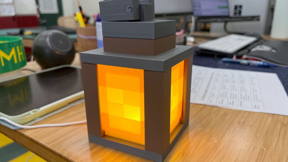 A glowing 3D printed lantern with brown and grey frame pieces and illuminated yellow panels, sitting on a classroom desk.