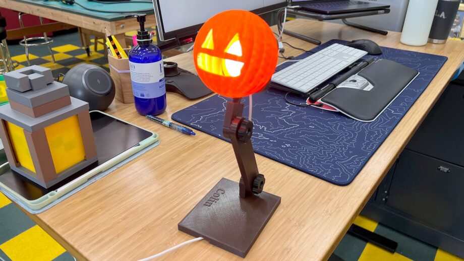 A 3D printed desk lamp with a glowing orange pumpkin-style light head and articulated brown arm, placed on a wooden desk alongside a tablet, keyboard, blue water bottle and a 3D printed lantern.