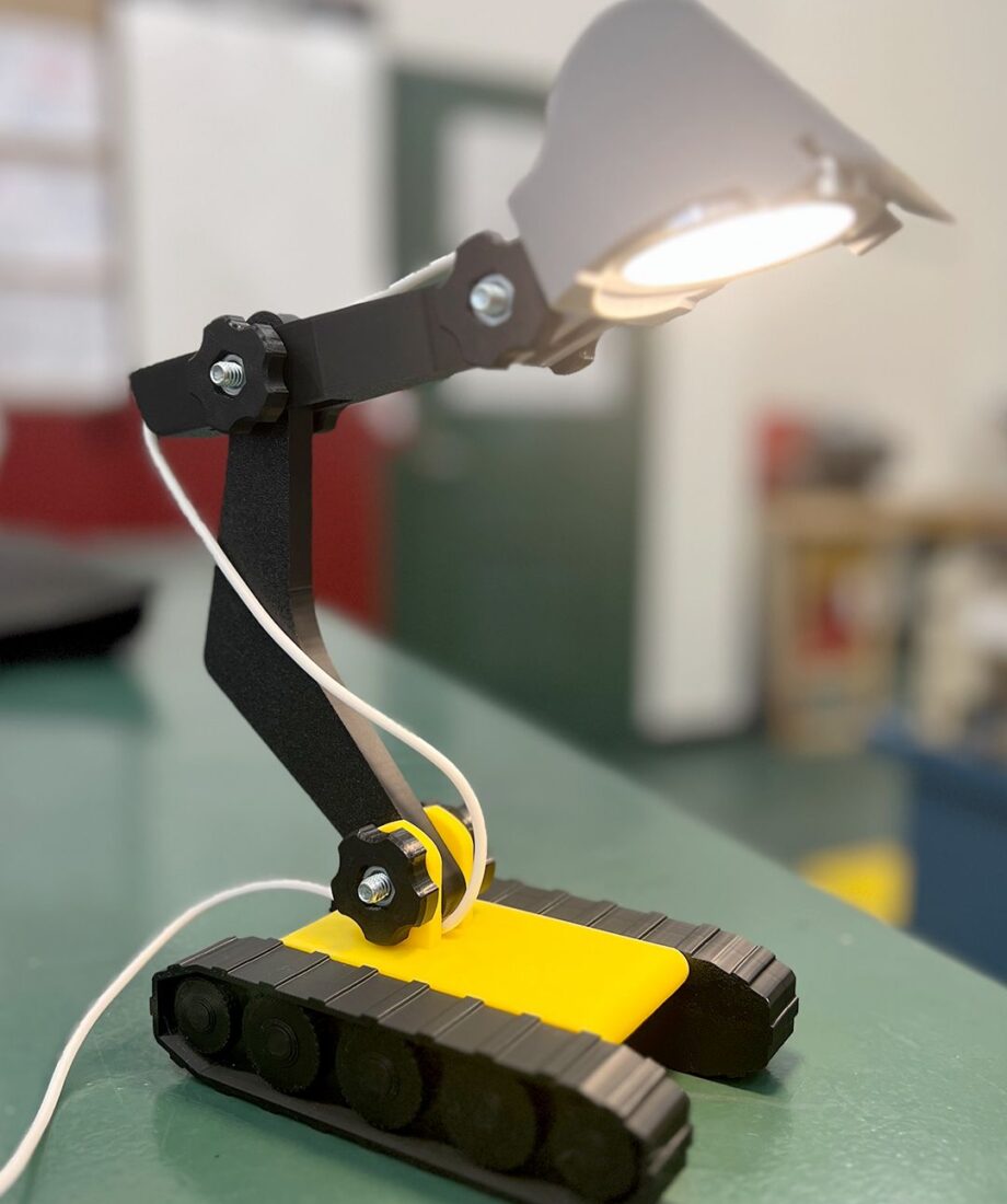A 3D printed desk lamp with articulated black and yellow mechanical arms and a lit lamp head, designed to resemble a small tracked robot.