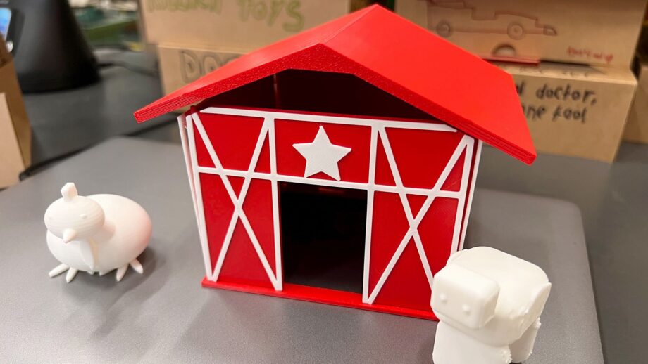 A small red 3D printed barn with white trim and a star above the doorway, shown on a tabletop with two white 3D printed animal figures beside it.