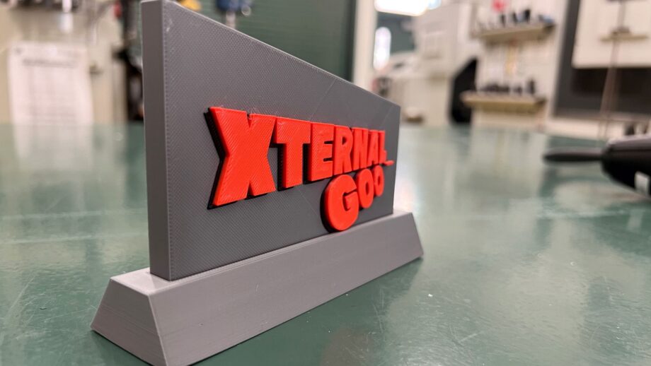 A grey 3D printed desk nameplate with raised red lettering that reads “Xternal Goo.”