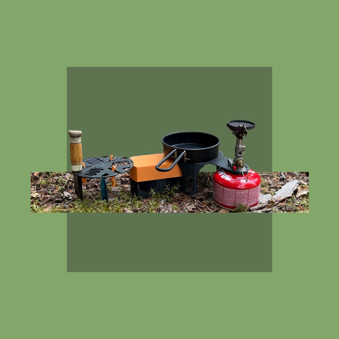 A compact 3D printed camping cooking station set up on forest ground. It includes a black stove platform attached to a red gas canister, a small frying pan, an orange table platform, and a side storage platform with a wooden-handled tool.