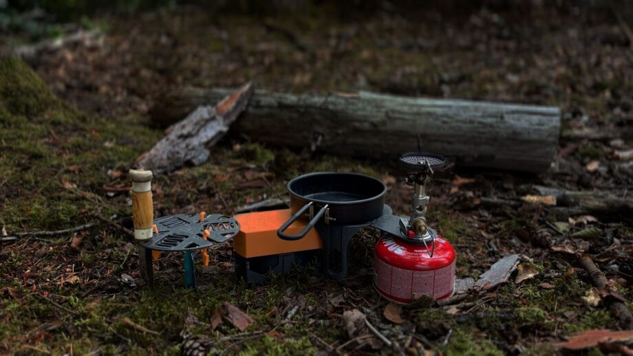 3D printed camp kitchen setup in a forest, including a compact stove stand, cookware, and a gas burner arranged on the ground.