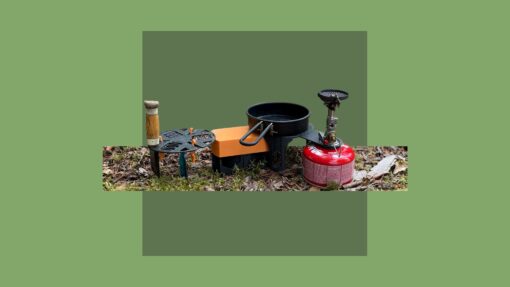 A compact 3D printed camping cooking station set up on forest ground. It includes a black stove platform attached to a red gas canister, a small frying pan, an orange table platform, and a side storage platform with a wooden-handled tool.