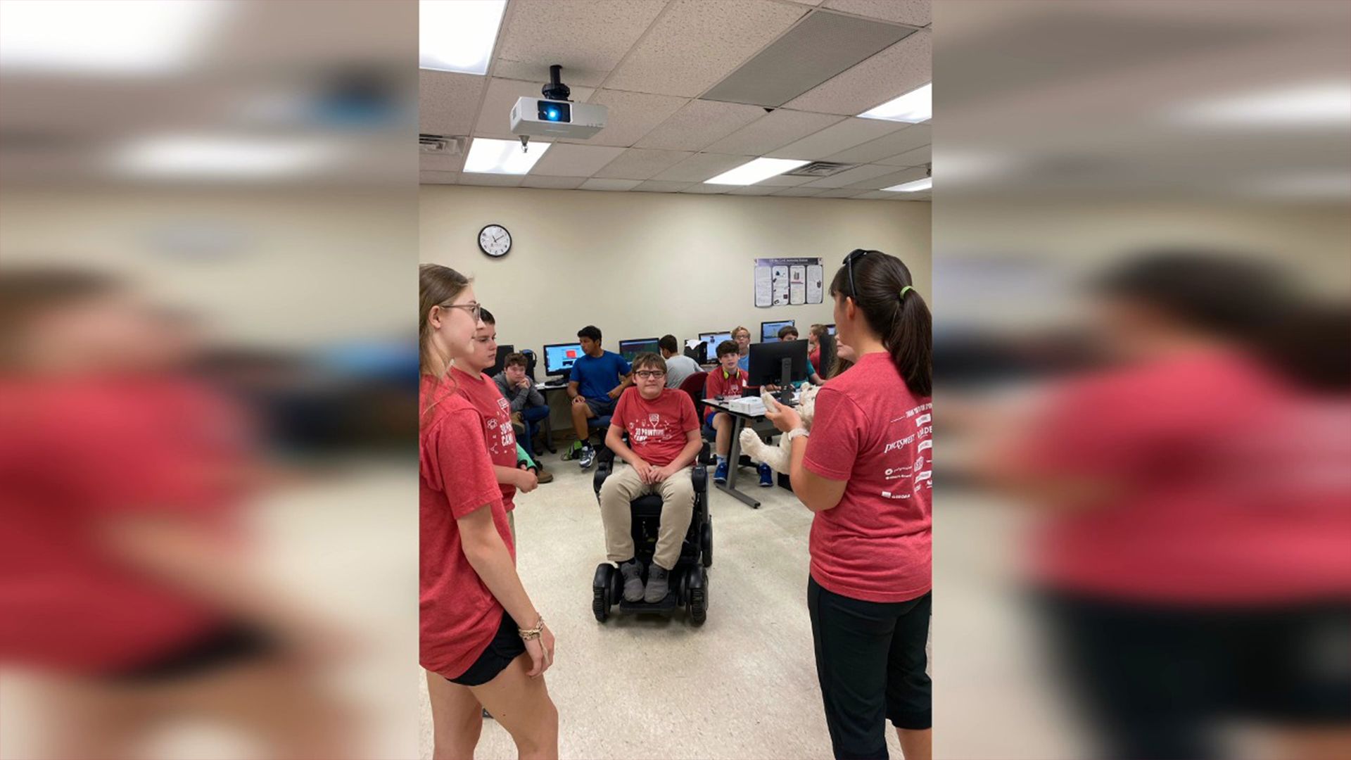 Students in a classroom gather around a student using a powered wheelchair while peers in matching red shirts lead a discussion, with computers and other students visible in the background.