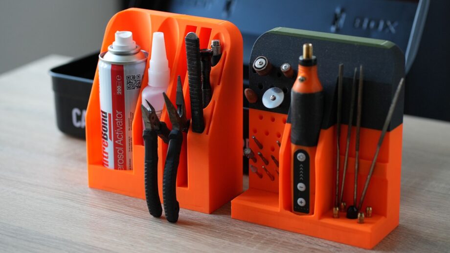 Two bright orange 3D printed organiser modules displayed on a desk, designed to hold glue, pliers, rotary tool bits, and hand tools in precise, custom-shaped slots.