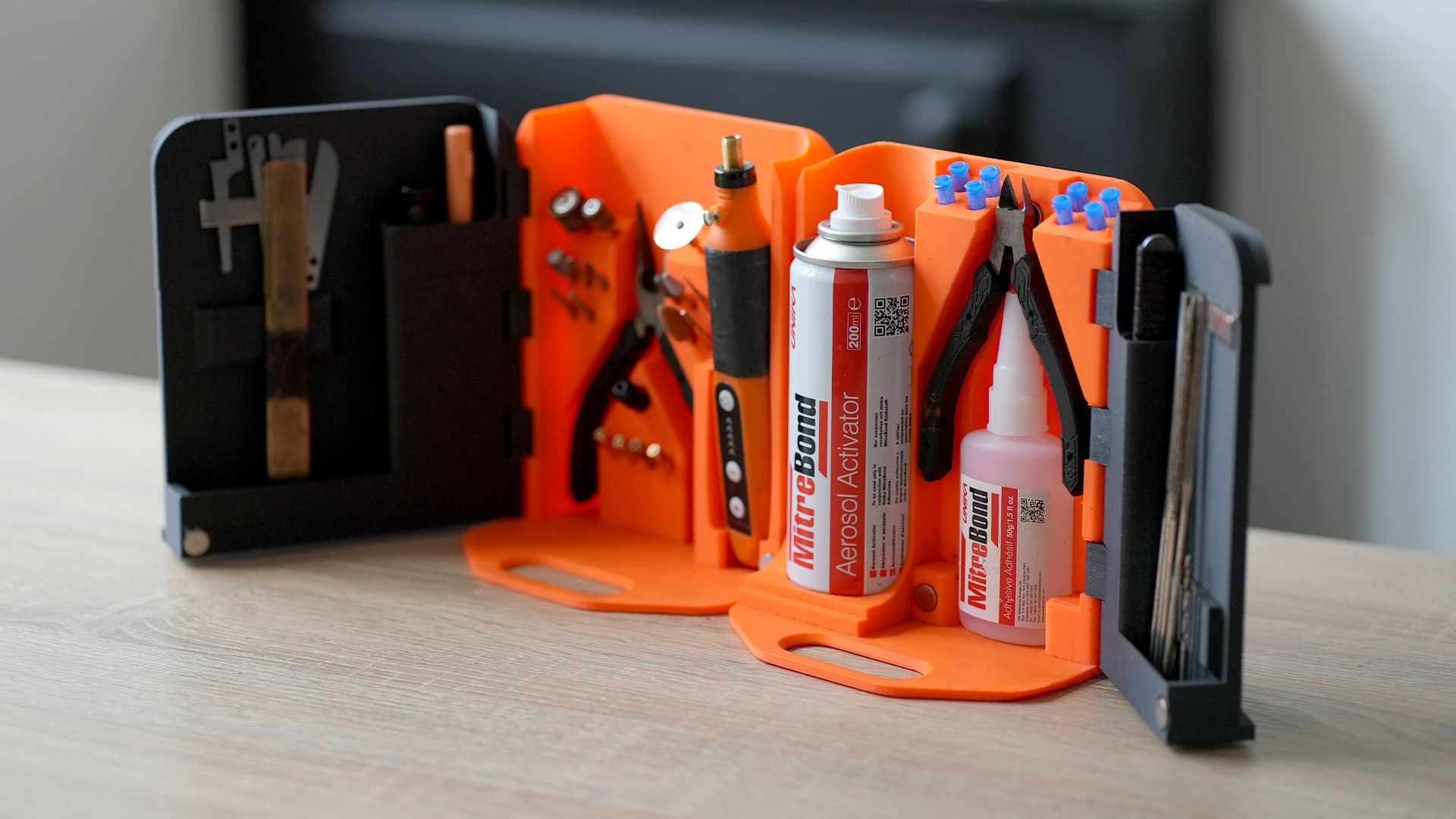 Advanced fold-out storage organiser with modular orange and black sections holding tools, adhesives, and accessories on a wooden table.