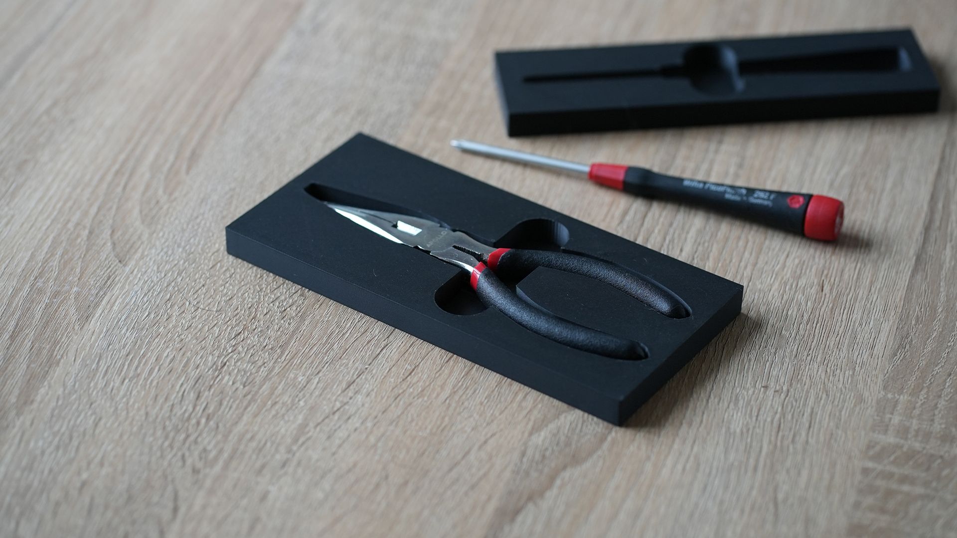 3D printed tool organiser insert holding needle-nose pliers, with a screwdriver placed nearby on a wooden desk.