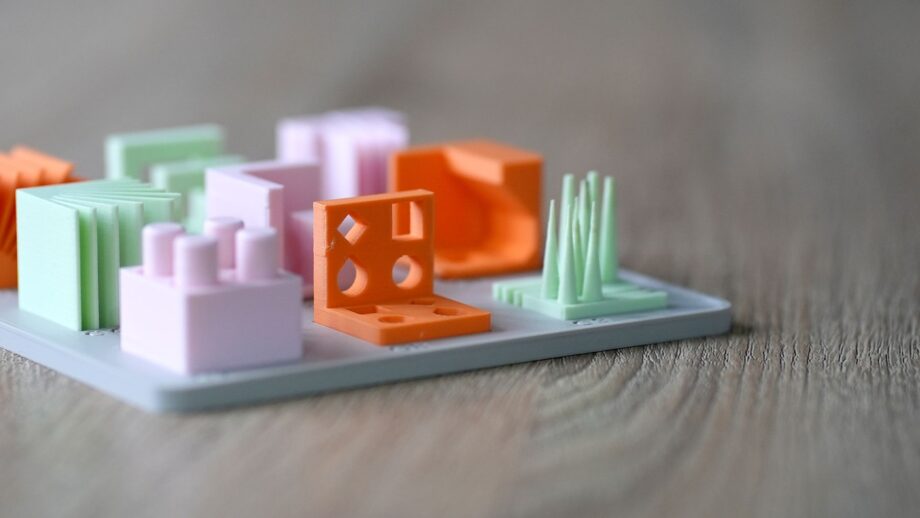 Close-up of a 3D printed design principles model set on a base plate, showing colourful test cubes with holes, overhangs, and spike features on a wooden surface.