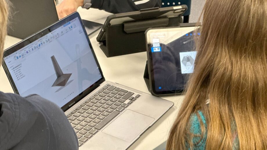 Students working on a spinning top design in Autodesk Fusion, with a tutorial displayed on a nearby tablet.