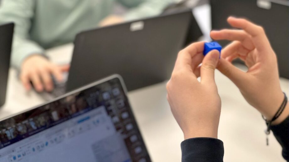 Student holding a small blue 3D printed calibration cube beside a laptop.